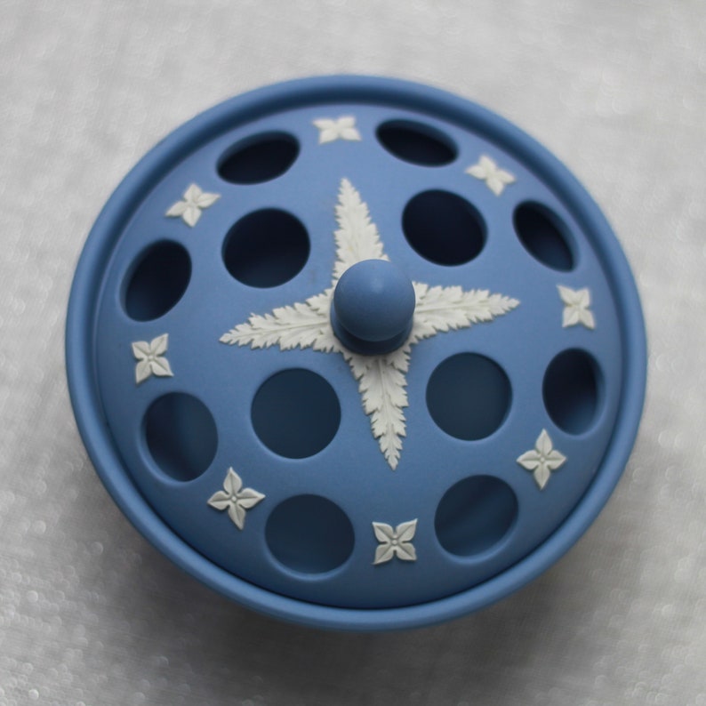 May include: A light blue ceramic container with a lid featuring a central starburst design and floral accents. The lid has circular openings and a small knob. The container is a decorative piece.