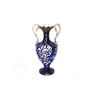 Può includere: Un vaso in ceramica blu scuro con disegni floreali bianchi e oro. Il vaso ha due manici e una base a piedistallo. Il bordo e le maniglie sono rifiniti in oro. Il motivo floreale è centrato sul vaso.