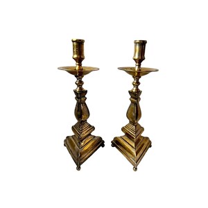 Può includere: Due candelabri antichi in ottone. Ogni candelabro presenta una base geometrica, uno stelo decorativo, un piattino e un portacandele cilindrico. I candelabri sono di un caldo colore dorato. Il design è dettagliato, su sfondo bianco.