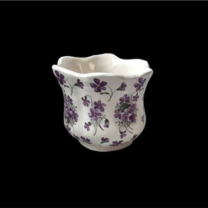 Könnte beinhalten: Ein weißer Keramik-Blumentopf mit gewelltem Rand, verziert mit einem sich wiederholenden Muster aus lila Veilchen und grünen Blättern. Das florale Design bedeckt die Außenseite und schafft ein dekoratives Stück.