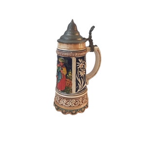 May include: A ceramic beer stein with a hinged metal lid. The stein features a detailed relief design with a woman in traditional clothing, floral accents, and a decorative handle. The colors include cream, blue, brown, and red.