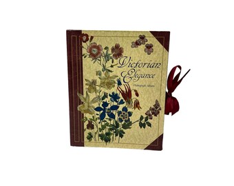 May include: A vintage photograph album with a floral design and the words "Victorian Elegance." The cover features a cream background with colorful flowers and a burgundy spine and corners. A burgundy ribbon is attached.