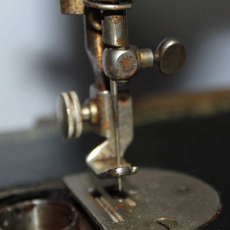May include: Close-up of an antique sewing machine needle and presser foot. The metal components show signs of age with rust and wear. The needle is positioned above a metal plate with feed dogs.