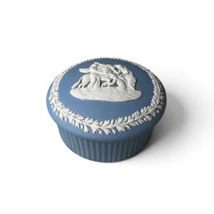 Può includere: Un portagioie rotondo in ceramica blu con un design in rilievo bianco sul coperchio. Il coperchio presenta una scena centrale con figure e un cavallo circondati da un bordo di foglie di alloro. La base della scatola ha una trama a coste.