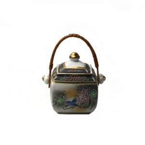 May include: A square ceramic container with a lid and a woven handle. The container is white with a black top and gold trim. A bird and floral design decorates the front. The handle is brown.