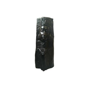 May include: A dark brown, carved wooden sculpture of a Native American woman's face and upper body. The sculpture features detailed carvings of a headdress, necklace, and facial features. The artwork is set against a white background.