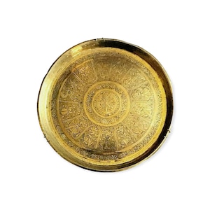 Op de afbeelding: Een ronde, goudkleurige metalen dienblad met ingewikkelde bloemen- en geometrische patronen die over het oppervlak zijn geëtst. De lade heeft een opstaande rand en een centraal cirkelvormig ontwerp, dat gedetailleerd vakmanschap laat zien. De algehele esthetiek is sierlijk en decoratief.