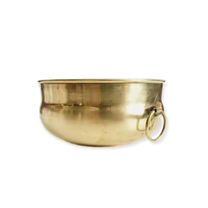 Pode incluir: Uma tigela de latão polido com corpo arredondado e uma alça circular. A borda da tigela é ligeiramente alargada. A superfície de latão reflete a luz. Ideal para decoração.