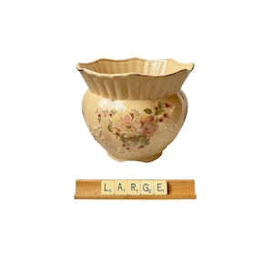 May include: A cream-colored ceramic planter with a scalloped rim and a gold-toned edge. The body features a floral pattern with pink and white flowers and green leaves. Below the planter, a wooden rack displays tiles spelling the word LARGE.