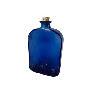 Può includere: Una bottiglia di vetro blu cobalto con tappo in sughero. La bottiglia ha una forma arrotondata con un fronte piatto e un motivo decorativo a onde sulla base. La bottiglia è vuota e ha un'estetica vintage.