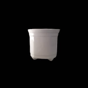 May include: A small, white ceramic plant pot with a glossy finish. The pot has a rectangular shape with rounded corners and sits on three small feet. The top edge has a slight inward curve. The background is solid black.