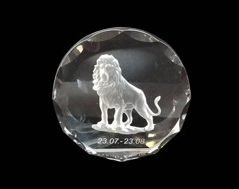 May include: A clear, faceted crystal paperweight featuring a detailed, white lion sculpture. The lion stands on a base with the dates "23.07.-23.08" etched below. The paperweight is round with a beveled edge.
