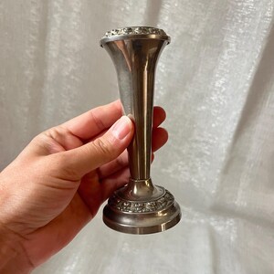 May include: A silver-toned bud vase with a flared top and a decorative base. The vase has a slender, elongated body and is likely made of metal. The top and base feature ornate detailing, adding to its vintage appeal. The vase is held by a hand.