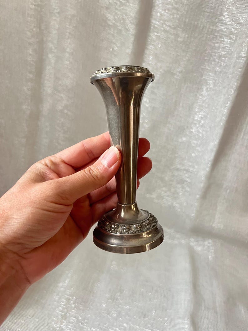 May include: A silver-toned bud vase with a flared top and a decorative base. The vase has a slender, elongated body and is likely made of metal. The top and base feature ornate detailing, adding to its vintage appeal. The vase is held by a hand.