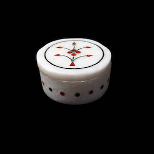 May include: A small, round white marble box with a lid. The lid features a floral design with red, orange, and turquoise accents. The box has a black border and is decorated with red and black dots around the base. The box is set against a black background.