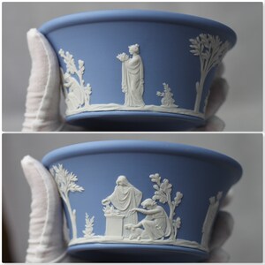 May include: A blue ceramic bowl with white bas-relief figures and foliage. The bowl has a wide rim and a slightly flared shape. The white relief depicts classical figures and trees, creating a decorative effect. The bowl is held by a gloved hand.