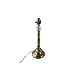 Może przedstawiać: Mała lampa stołowa w stylu antycznym z wykończeniem ze szczotkowanego mosiądzu. Lampa posiada przezroczysty kryształowy akcent i standardowe gniazdo żarówki. Podstawa jest okrągła, a przewód zasilający wychodzi od dołu.