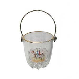 Puede incluir: Cubeta de hielo de vidrio transparente con borde dorado y asa de metal retorcido. La cubeta presenta una imagen decorativa de dos personas montando a caballo. La base tiene una serie de protuberancias redondeadas.