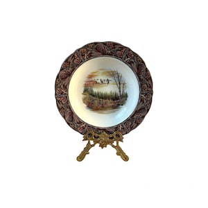 May include: A decorative plate with a nature scene in the centre, featuring a landscape with trees and birds. The plate has a brown, textured border with pinecones and is displayed on a gold-coloured stand.