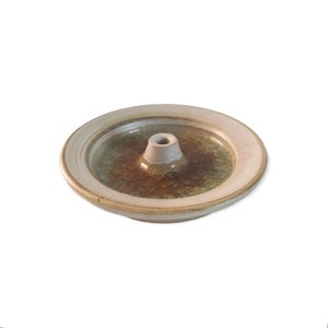 May include: A ceramic incense holder with a circular design. The holder features a central cone for incense and a surrounding dish. The color palette includes beige, brown, and green tones, creating a natural aesthetic. The holder is designed to catch ash.