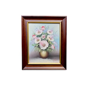 May include: A framed floral oil painting featuring a bouquet of pink and white flowers in a gold vase. The painting has a light blue background and is encased in a dark brown wooden frame with a gold inner border. The artwork is a still life.