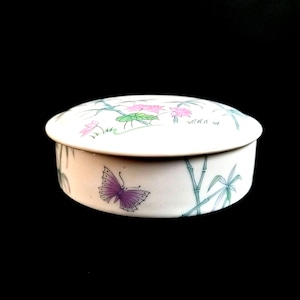 Puede incluir: Caja de baratijas de cerámica blanca con tapa, con una mariposa morada y bambú. La tapa está decorada con flores rosas y hojas verdes. La caja redonda está sobre un fondo negro.