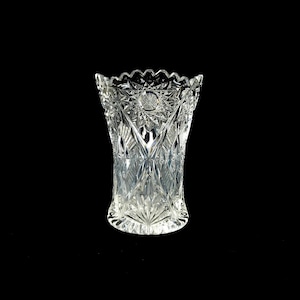 May include: A clear, cut-glass vase with a scalloped rim. The vase features intricate geometric patterns and a starburst design at the top. The vase is approximately 18 cm tall and has a wide base.