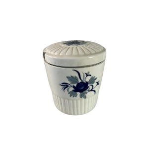 May include: A white ceramic jar with a lid, decorated with a blue floral pattern. The jar has a ribbed texture on the lower part and the lid. The floral design includes blue roses and leaves. A small spoon is on the side of the jar.