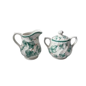 May include: A white ceramic creamer and sugar bowl set. The set features a green ivy leaf design with a scalloped rim. The sugar bowl has a lid with a matching ivy leaf pattern. The creamer has a curved handle and spout.