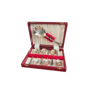 May include: A collection of silver spoons presented in a red, hinged box. The spoons feature ornate, shell-shaped bowls and are displayed on a white satin lining. The box is open, showcasing the spoons and a red velvet holder.