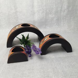 K&ouml;nnte beinhalten: Drei dunkelbraune Kerzenhalter aus Holz in verschiedenen Gr&ouml;&szlig;en. Jeder hat eine halbkreisf&ouml;rmige Form mit einem Loch f&uuml;r eine Kerze. Ein Band aus rosa und goldenen Blumenmustern verziert die Oberseite jedes Halters. Ein Zweig Lavendel und Gr&uuml;nzeug ist in der N&auml;he der Halter platziert.