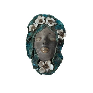 Może przedstawiać: Ceramiczna rzeźba twarzy o spokojnym wyrazie. Twarz jest stonowana szara, z turkusowymi włosami i białymi kwiatowymi akcentami. Rzeźba ma gładkie wykończenie i jest umieszczona na białym tle.