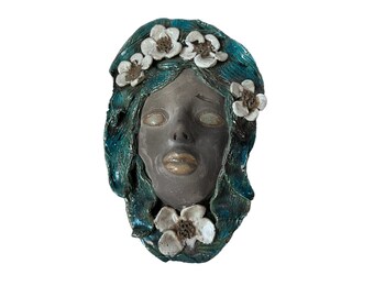 May include: A ceramic face sculpture with a serene expression. The face is a muted gray, with teal-colored hair and white flower accents. The sculpture has a smooth finish and is set against a white background.