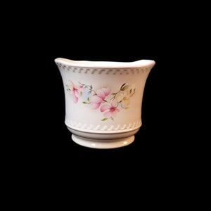 May include: White ceramic planter with a flared shape, featuring a delicate floral design of pink and white blossoms with green leaves. The planter has a decorative beaded rim and a grey base.