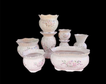 May include: A collection of cream-colored ceramic flower pots and vases. The pots and vases have a ruffled edge and are decorated with floral patterns in pink, green, and blue. The collection includes various sizes and shapes.
