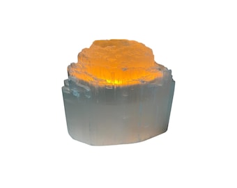 May include: A light blue selenite crystal candle holder with a warm, glowing light emanating from within. The crystal is cylindrical with a textured, layered appearance. The top has a circular opening for a candle.