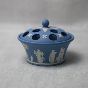 May include: A blue ceramic flower frog with a lid featuring a knob and several holes. The sides of the frog are decorated with white relief figures and floral designs. A classic piece for floral arrangements.
