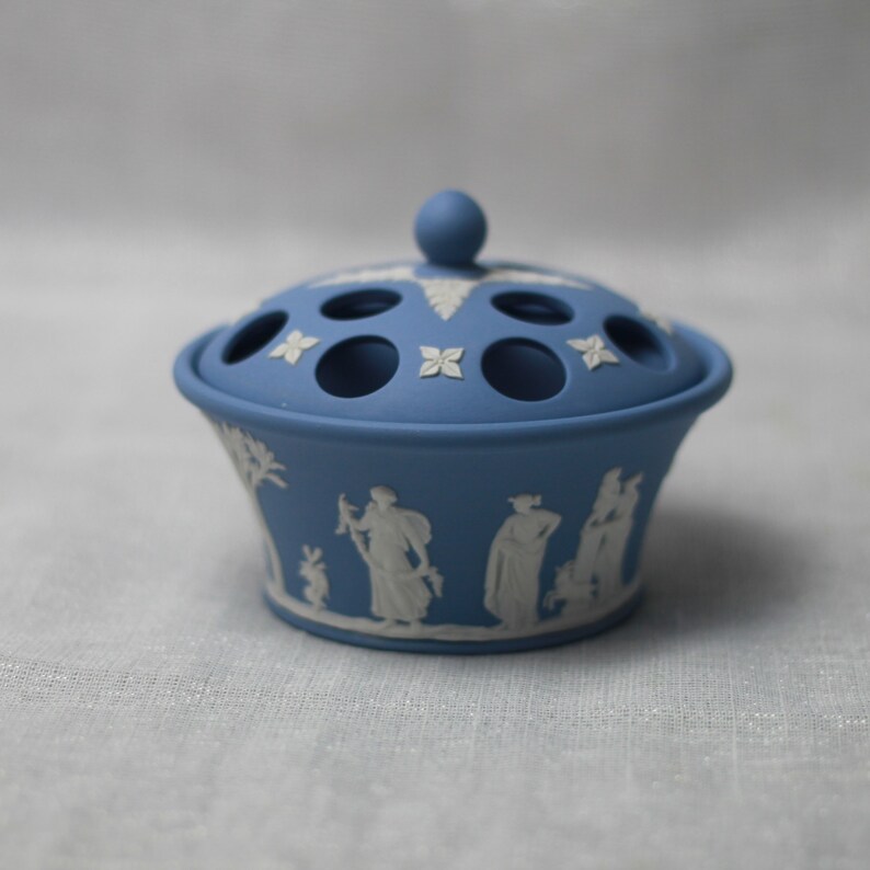 May include: A blue ceramic flower frog with a lid featuring a knob and several holes. The sides of the frog are decorated with white relief figures and floral designs. A classic piece for floral arrangements.