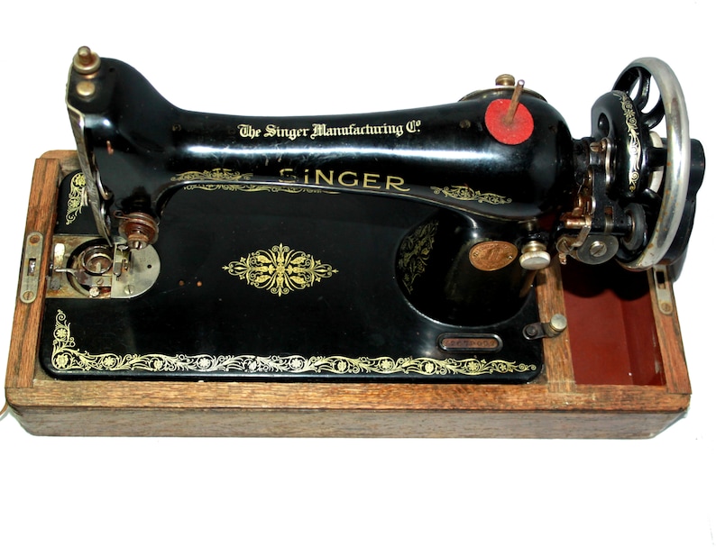 May include: A vintage Singer sewing machine, black with gold accents, mounted on a wooden base. The machine features the Singer Manufacturing Co. logo and decorative floral patterns. The hand wheel is visible on the right side.