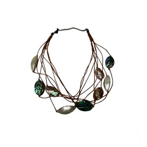 May include: A multi-strand necklace featuring oval-shaped iridescent beads in shades of green, silver, and brown. The beads are strung on brown cords and connected with a chain clasp. The necklace is set against a white background.