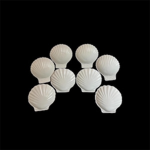 Könnte beinhalten: Ein Set aus acht weißen Keramikschalen in Form von Jakobsmuscheln. Jede Schale weist ein fächerartiges Design mit strahlenförmigen Rillen auf, die von der Basis bis zum gewellten Rand verlaufen und ein einheitliches Erscheinungsbild erzeugen.
