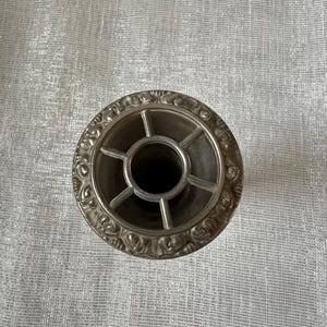 May include: A silver-colored, circular object with an ornate, floral-patterned rim. The center features a cross-shaped design with a dark interior. The object appears to be a decorative piece, possibly for a table setting or home decor.