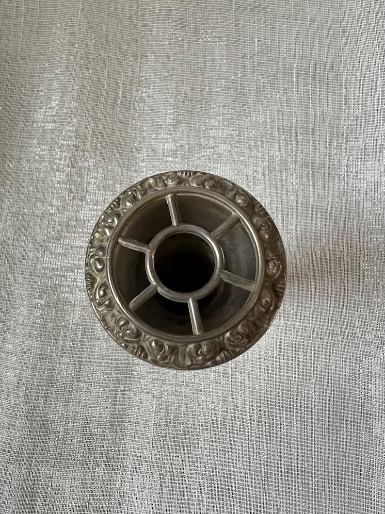 May include: A silver-colored, circular object with an ornate, floral-patterned rim. The center features a cross-shaped design with a dark interior. The object appears to be a decorative piece, possibly for a table setting or home decor.