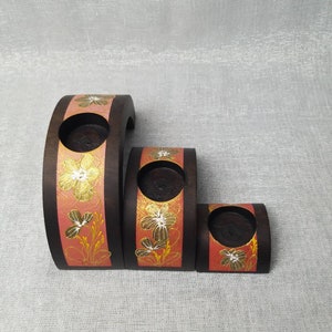 K&ouml;nnte beinhalten: Set aus drei unterschiedlich gro&szlig;en Kerzenhaltern aus Holz. Die dunkelbraunen Halterungen weisen ein bandf&ouml;rmiges Element aus korallenfarbener Farbe mit goldenen Blumendesigns auf. Jeder Halter hat eine runde &Ouml;ffnung f&uuml;r eine Kerze.
