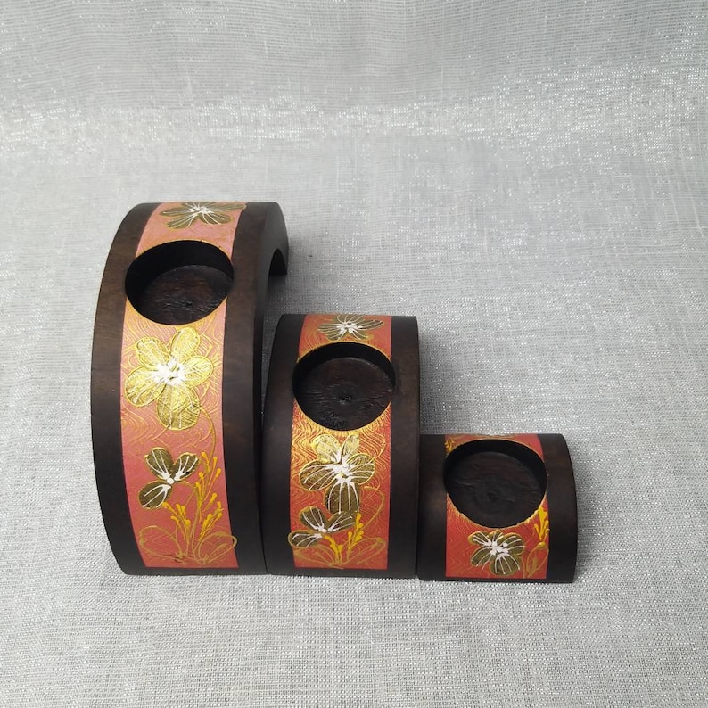 K&ouml;nnte beinhalten: Set aus drei unterschiedlich gro&szlig;en Kerzenhaltern aus Holz. Die dunkelbraunen Halterungen weisen ein bandf&ouml;rmiges Element aus korallenfarbener Farbe mit goldenen Blumendesigns auf. Jeder Halter hat eine runde &Ouml;ffnung f&uuml;r eine Kerze.