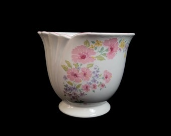 May include: A white ceramic planter with a scalloped rim and a pedestal base. The planter is decorated with pink, purple, and yellow floral patterns. The background is solid black.