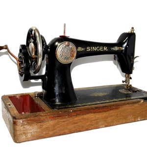 May include: A vintage Singer sewing machine, black with gold accents, set on a wooden base. The machine features a hand crank, a large balance wheel, and intricate detailing. The word "SINGER" is visible on the arm of the machine.