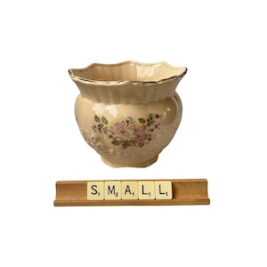 Puede incluir: Jardinera de cerámica color crema con borde festoneado y diseño floral. La jardinera tiene un ribete dorado y detalles en relieve. La palabra "SMALL" está escrita con fichas de letras en un soporte de madera.