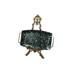 May include: A rectangular serving tray with a dark, speckled surface, supported by an ornate, golden metal stand. The tray has a handle on each side. The stand features decorative flourishes.