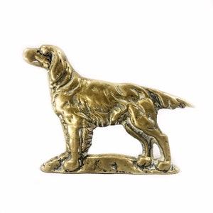 May include: A brass dog ornament in a standing position. The sculpture has a detailed texture and a golden colour. The dog's features are well-defined, with a pointed nose and a slightly raised tail. The ornament is on a small base.